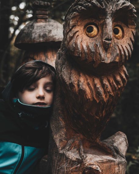 Ein Junge lehnt an einer Holzfigur einer Eule im Wald.
