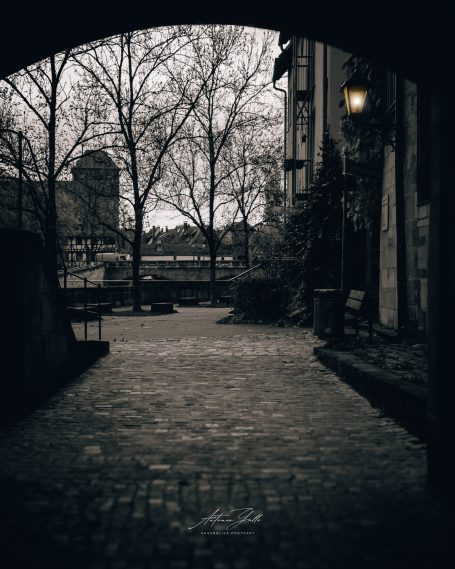 Dunkler Weg unter einem Bogen, umgeben von Bäumen und einer laterne, im Hintergrund Wasser.